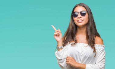 Kameraya bakarak yan el ve parmak ile işaret yüzünde kocaman bir gülümseme ile izole arka plan üzerinde güneş gözlüğü takmış genç güzel Arap kadın.