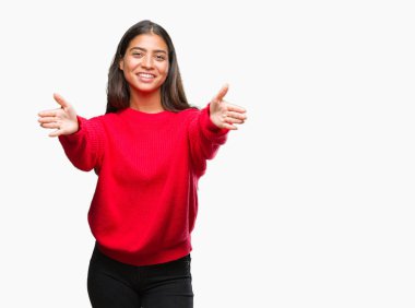 Sarılmak için kollarını açarak gülümseyerek kameraya bakarak izole arka plan üzerinde kış kazak giyen genç güzel Arap kadın. Neşeli ifade kucaklayan mutluluk.