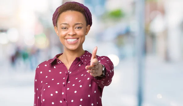 Beautiful young african american woman wearing head scarf over isolated background smiling friendly offering handshake as greeting and welcoming. Successful business.