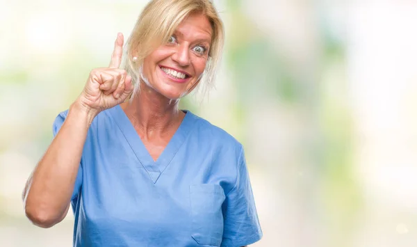 Middle age blonde woman wearing doctor nurse uniform over isolated background pointing finger up with successful idea. Exited and happy. Number one.