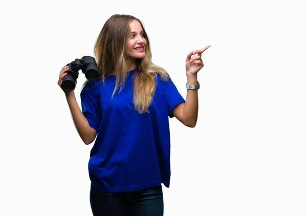 年轻美丽的金发女子寻找通过双筒望远镜在孤立的背景非常愉快的指向与手和手指的一侧 — 图库照片