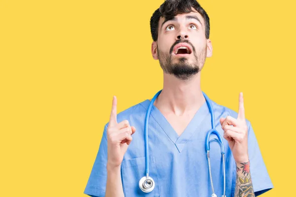 Young handsome nurse man wearing surgeon uniform over isolated background amazed and surprised looking up and pointing with fingers and raised arms.