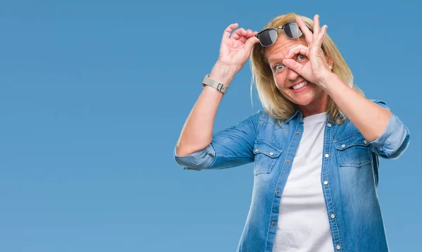 中年金发碧眼的女人戴着太阳镜在孤立的背景做确定的手势与手微笑 眼睛通过手指看着快乐的脸 — 图库照片