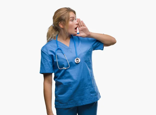 年轻的高加索医生妇女穿着外科医生制服在孤立的背景大喊和尖叫的一边 手放在嘴 沟通理念 — 图库照片