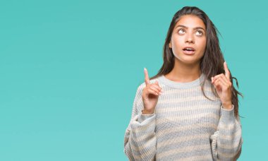 İzole arka plan üzerinde kış kazak giyen genç güzel Arap kadın hayrete ve yukarıya bakıyor ve parmakları ile işaret ve silah kaldırdı şaşırttı.