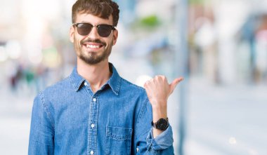 Yakışıklı delikanlı arıyor ve başparmak yanında yukarıyı mutlu yüz ile gülümseyen izole arka plan üzerinde güneş gözlüğü takıyor.