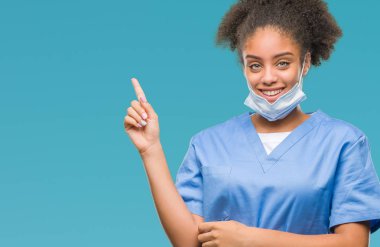 Kameraya bakarak yan el ve parmak ile işaret eden genç afro Amerikalı doktor kadın yüzünde kocaman bir gülümseme ile izole arka plan üzerinde.