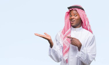 Geleneksel keffiyeh hayret ve el ile sunulması ve parmak ile işaret ederken kameraya gülümseyen izole arka plan üzerinde giyen genç Arapça Afrika adam.