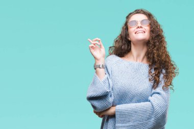 Kameraya bakarak yan el ve parmak ile işaret yüzünde kocaman bir gülümseme ile izole arka plan üzerinde güneş gözlüğü takıyor güzel esmer kıvırcık saçlı Genç kız.