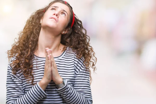 Beautiful brunette curly hair young girl wearing stripes sweater over isolated background begging and praying with hands together with hope expression on face very emotional and worried. Asking for forgiveness. Religion concept.