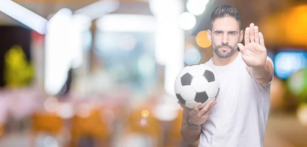Young man holding soccer football ball over isolated background with open hand doing stop sign with serious and confident expression, defense gesture