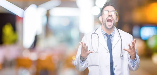 Handsome young doctor man over isolated background crazy and mad shouting and yelling with aggressive expression and arms raised. Frustration concept.