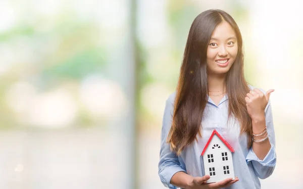 Young asian woman real state agent holding house isolated background pointing and showing with thumb up to the side with happy face smiling