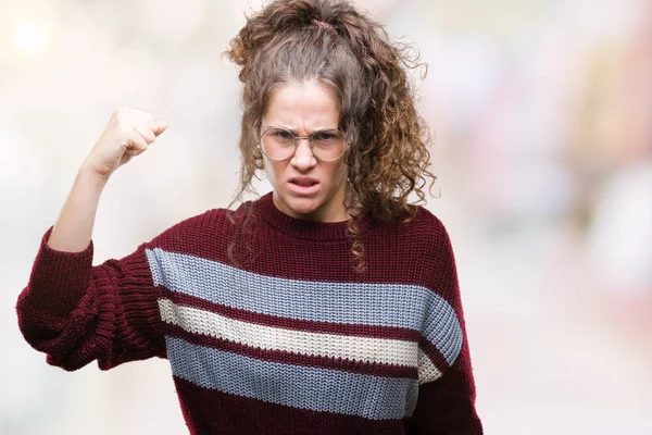 Beautiful brunette curly hair young girl wearing glasses over isolated background angry and mad raising fist frustrated and furious while shouting with anger. Rage and aggressive concept.