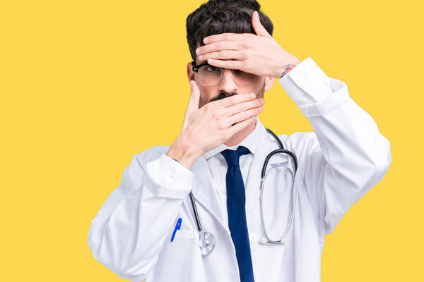Young doctor man wearing hospital coat over isolated background Covering eyes and mouth with hands, surprised and shocked. Hiding emotion