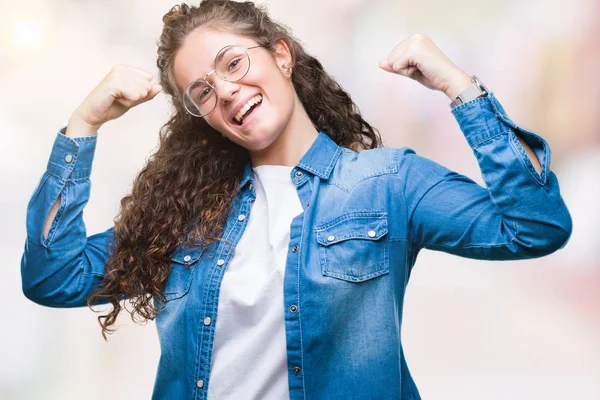 Beautiful young brunette curly hair girl wearing glasses over isolated background very happy and excited doing winner gesture with arms raised, smiling and screaming for success. Celebration concept.