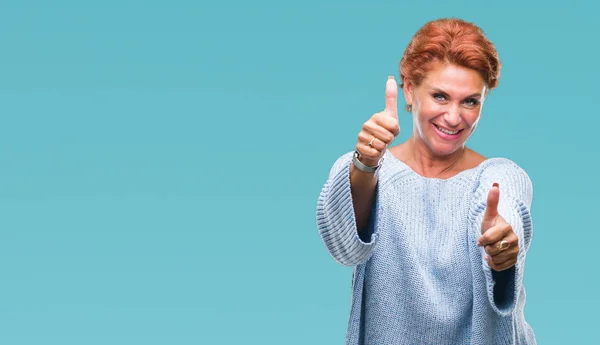 Atrractive senior caucasian redhead woman wearing winter sweater over isolated background approving doing positive gesture with hand, thumbs up smiling and happy for success. Looking at the camera, winner gesture.