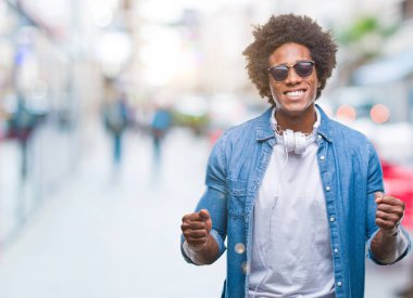 Üzerinden müzik kulaklık giyen Afro Amerikalı adam arka plan çok mutlu ve heyecanlı kaldırdı, silah ile kazanan jest yapıyor gülümseyen ve başarı için çığlık izole. Kutlama kavramı.