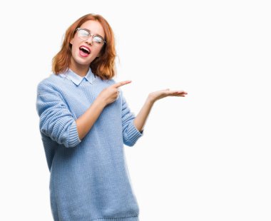 İzole arka plan giyen kış kazak hayret ve el ile sunulması ve parmak ile işaret ederken kameraya gülümseyen genç güzel kadın.