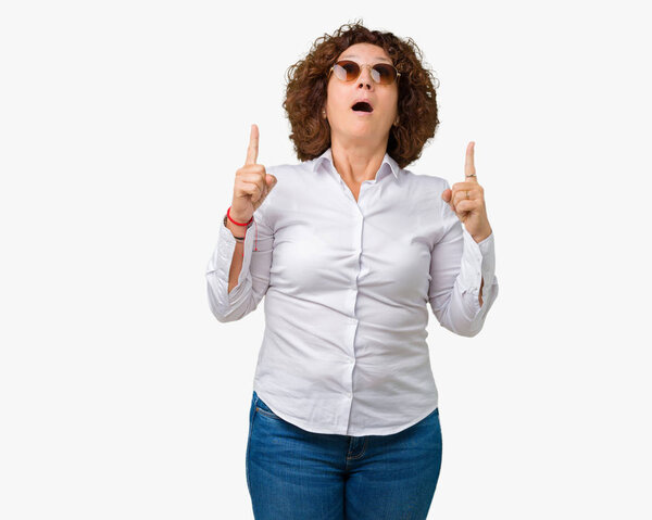 Beautiful middle ager senior business woman wearing sunglasses over isolated background amazed and surprised looking up and pointing with fingers and raised arms.