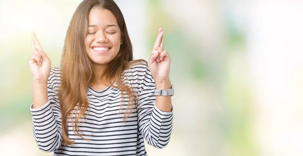 年轻美丽的黑发女人穿着条纹毛衣在孤立的背景微笑交叉手指与希望和眼睛闭上 运气和迷信的概念 — 图库照片