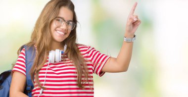 Yüzünde kocaman bir gülümseme ile izole arka plan üzerinde kulaklık ve sırt çantası giyen genç güzel esmer öğrenci kadın kameraya bakarak yan el ve parmak ile işaret.