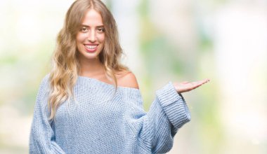 Neşeli sunulması ve kameraya bakarak avucunun ile işaret gülümseyen izole arka plan üzerinde kış kazak giyen güzel genç sarışın kadın.