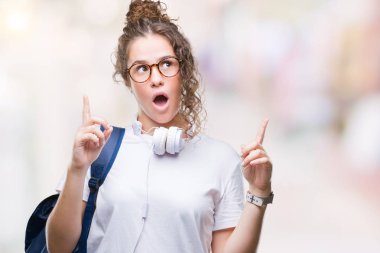 Sırt çantası ve kulaklık izole arka plan üzerinde giyen genç esmer öğrenci kız hayran ve yukarıya bakıp parmakları ile işaret şaşırttı ve silah kaldırdı.