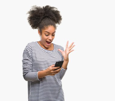 Genç afro Amerikalı kadın manifatura üzerinden Smartphone kullanarak büyük gülümseme ile çığlık zaferini kutluyor arka plan çok mutlu ve heyecanlı, kazanan ifade izole ve ellerini kaldırdı