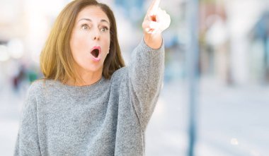 Güzel orta yaş kadın giyiyor kış kazak izole arka plan ile sürpriz devam et, parmakla üzerinde açık ağız ifade, önünde bir şey hayret