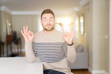 Evde bir kazak giyen genç yakışıklı adam rahatla ve gülümseyen gözlerle kapalı parmak jestle meditasyon yapıyor. Yoga kavramı.