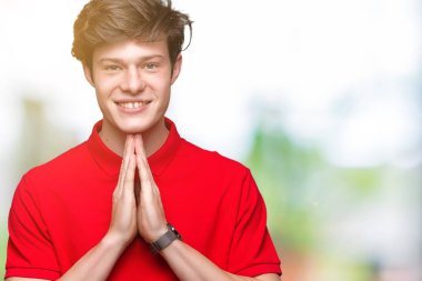 Birlikte emin gülümseyen bağışlanmak ellerle dua izole arka plan üzerinde kırmızı t-shirt giyen genç yakışıklı adam.