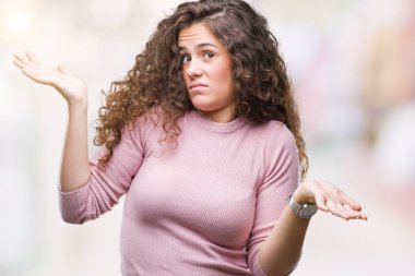 Güzel esmer kıvırcık saçlı Genç kız üzerinde pembe kazak giyiyor arka plan clueless ve şaşkın bir ifadeyle kollar ve eller kaldırdı izole. Şüphe kavramı.