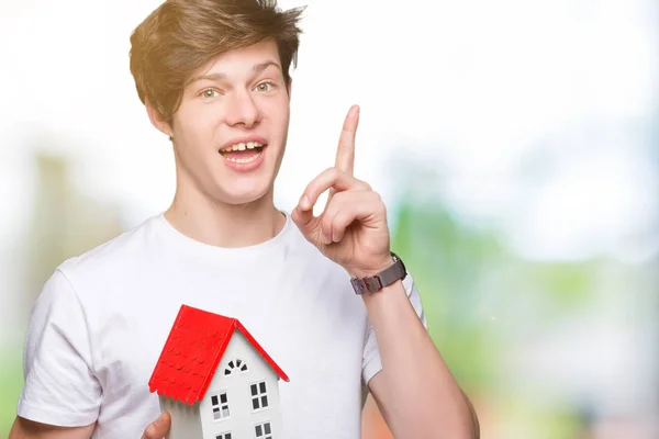 Young man holding house over isolated background surprised with an idea or question pointing finger with happy face, number one
