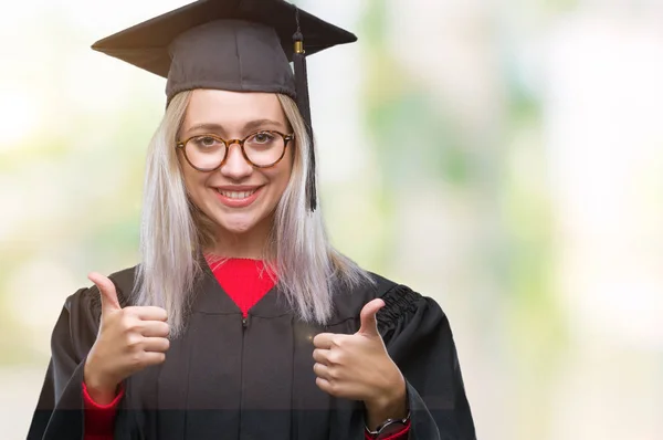 Young blonde woman wearing graduate uniform over isolated background approving doing positive gesture with hand, thumbs up smiling and happy for success. Looking at the camera, winner gesture.