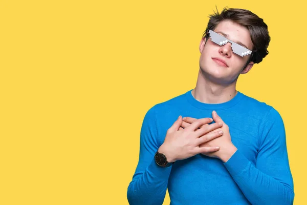 Young man wearing funny thug life glasses over isolated background smiling with hands on chest with closed eyes and grateful gesture on face. Health concept.