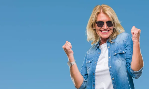 Middle age blonde woman wearing sunglasses over isolated background celebrating surprised and amazed for success with arms raised and open eyes. Winner concept.