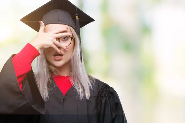 恥ずかしそうな顔で指を通して見る顔と手で目を覆うショックでピーク分離の背景の上の大学院の制服を着て若い金髪女性 — ストック写真
