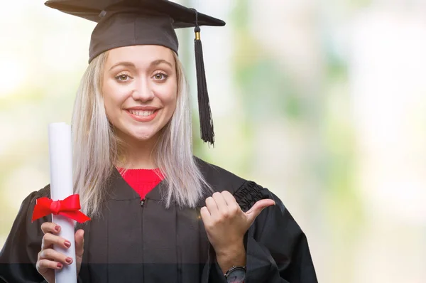 指していると幸せそうな顔を笑顔で側まで親指で示す分離の背景上の学位を保持する大学院の制服を着て若いブロンドの女性 — ストック写真