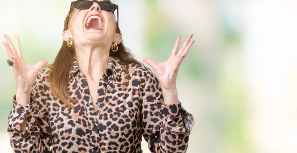 Middle age mature rich woman wearing sunglasses and leopard dress over isolated background crazy and mad shouting and yelling with aggressive expression and arms raised. Frustration concept.