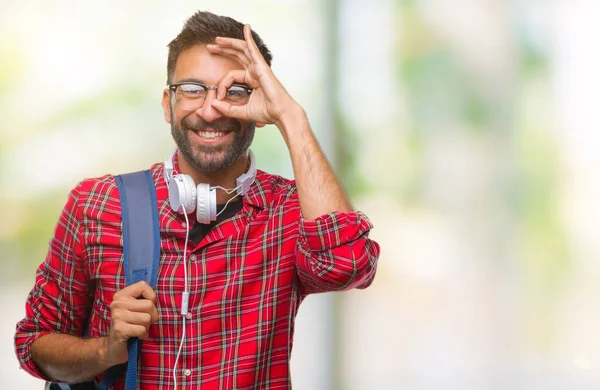 Gülümseyerek el parmakları ile mutlu yüz üzerinden seyir göz ile ok jest yapıyor izole arka plan üzerinde kulaklık ve sırt çantası giyiyor yetişkin İspanyol öğrenci adam.