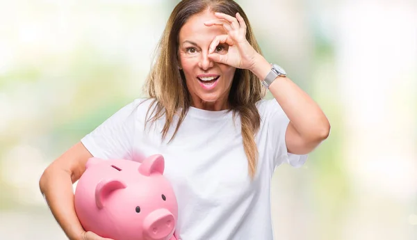 中年のヒスパニック系女性指を通して見る目に手で サインをして笑っている幸せそうな顔で孤立した背景に貯金箱を使ってお金を節約 — ストック写真