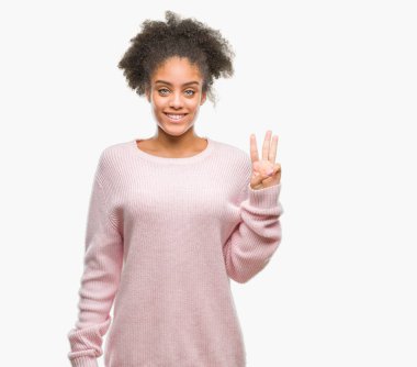 Genç afro Amerikalı kadın giyiyor kış kazak gösterilen ve parmakları ile işaret izole arka plan üzerinde üçüncü ise kendinden emin ve mutlu gülümseyerek.