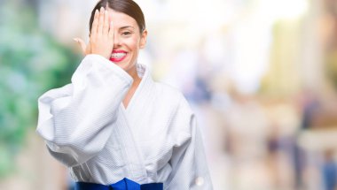 El yüz ve sürpriz duygu kendinden emin gülümseme ile tek gözüyle kapsayan izole arka plan üzerinde karate kimono üniforma giyen genç güzel kadın.