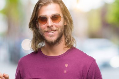 Kameraya bakarak yan el ve parmak ile işaret eden genç yakışıklı adam uzun saçlı yüzünde kocaman bir gülümseme ile izole arka plan üzerinde güneş gözlüğü takıyor.