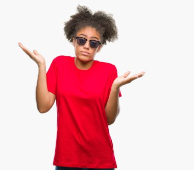 Genç afro Amerikalı kadın üzerinde güneş gözlüğü takıyor arka plan clueless ve şaşkın bir ifadeyle kollar ve eller kaldırdı izole. Şüphe kavramı.