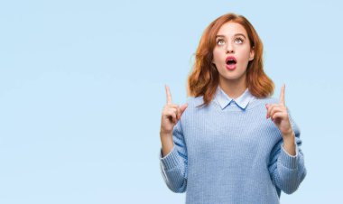 İzole arka plan kış kazak giyen genç güzel kadın hayrete ve yukarıya bakıyor ve parmakları ile işaret ve silah kaldırdı şaşırttı.