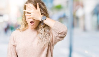 Mahcup ifadeyle parmaklarımın arasından seyir sweatershirt yüz ve gözler el ile kapsayan şok bakmak izole arka plan üzerinde giyen güzel sarışın genç kadın.