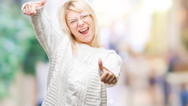 Kış kazak ve gözlük el ile pozitif jest yapıyor onaylama izole arka plan üzerinde giyen genç güzel sarışın kadın başparmak gülümseyen ve başarı için mutlu. Kamera, kazanan jest arıyorsunuz.