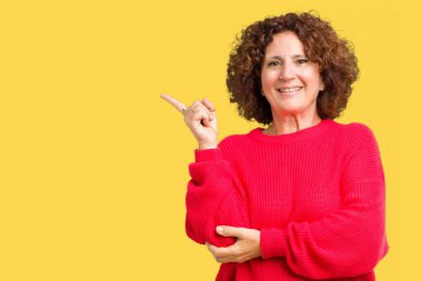 Güzel orta ager üst düzey kadın kırmızı kış kazak kameraya bakarak yan el ve parmak ile işaret yüzünde kocaman bir gülümseme ile izole arka plan üzerinde.